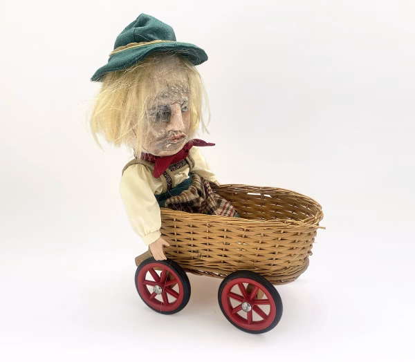Sculpture: Otto in wicker cart Hacked doll with soft body and china hands and feet. Doll face is replaced with uncanny head made from textile.
