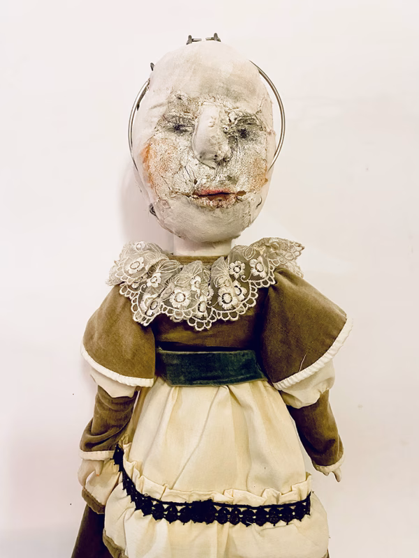 A doll wearing a sage green velvet and cream cotton dress. With a kilner jar clip over its head.