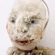 A doll wearing a sage green velvet and cream cotton dress. With a kilner jar clip over its head. Head view close-up.