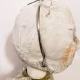 A doll wearing a sage green velvet and cream cotton dress. With a kilner jar clip over its head. Head view close-up.