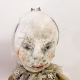 A doll wearing a sage green velvet and cream cotton dress. With a kilner jar clip over its head.