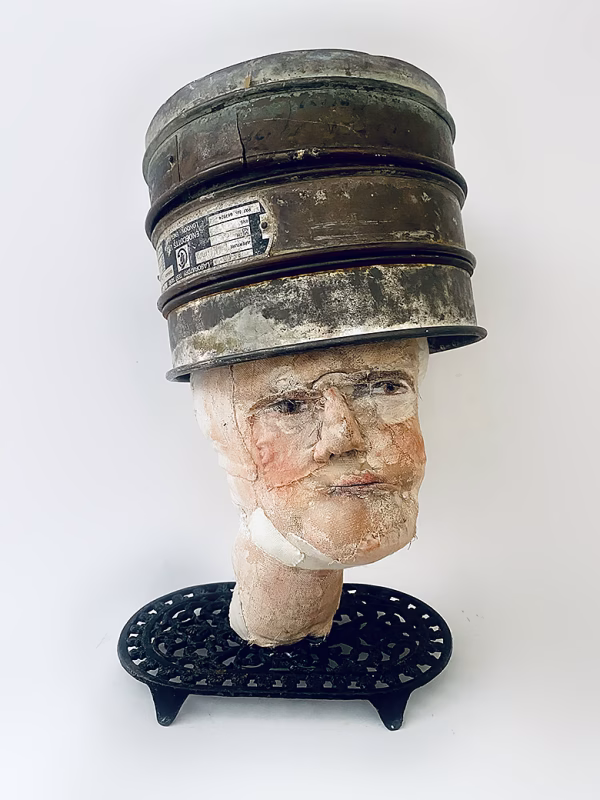 Head sculpture resting on a household iron trivet with a head piece of three stacked laboratory sieves.