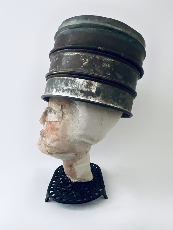 Head sculpture resting on a household iron trivet with a head piece of three stacked laboratory sieves.