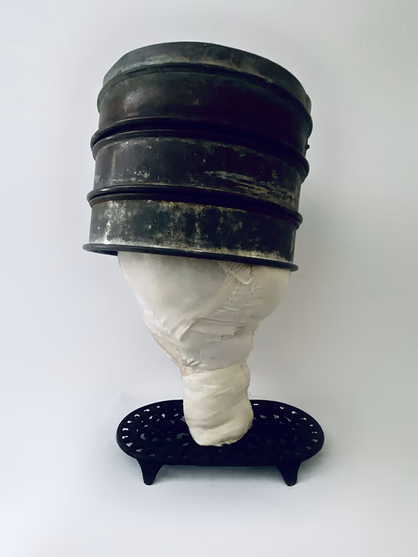 Head sculpture resting on a household iron trivet with a head piece of three stacked laboratory sieves.