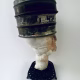 Head sculpture resting on a household iron trivet with a head piece of three stacked laboratory sieves.