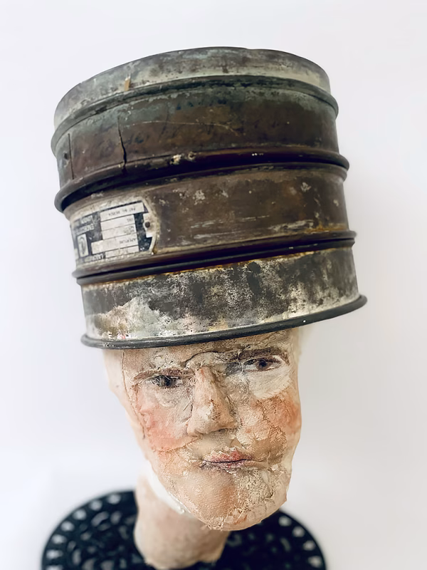 Head sculpture resting on a household iron trivet with a head piece of three stacked laboratory sieves.