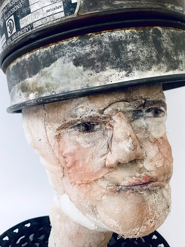 Close-up of head sculpture resting on a household iron trivet with a head piece of three stacked laboratory sieves.
