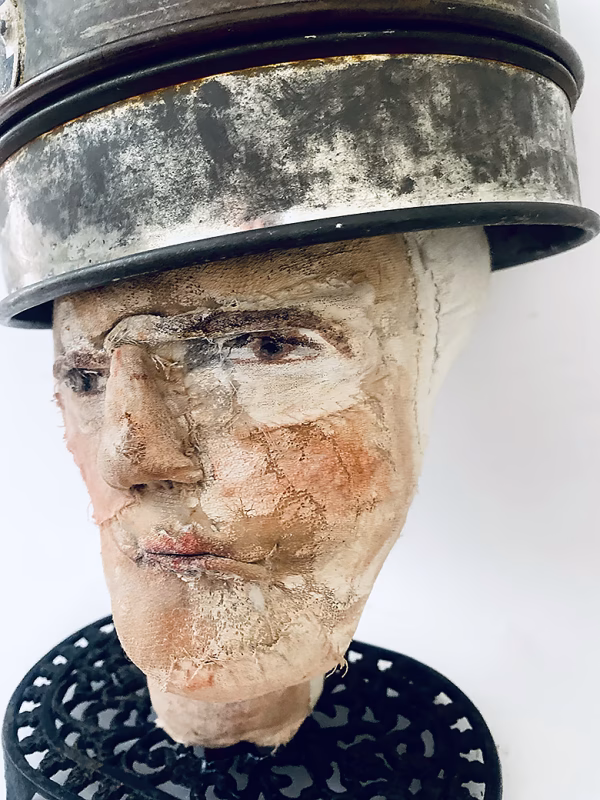 Close-up of head sculpture resting on a household iron trivet with a head piece of three stacked laboratory sieves.