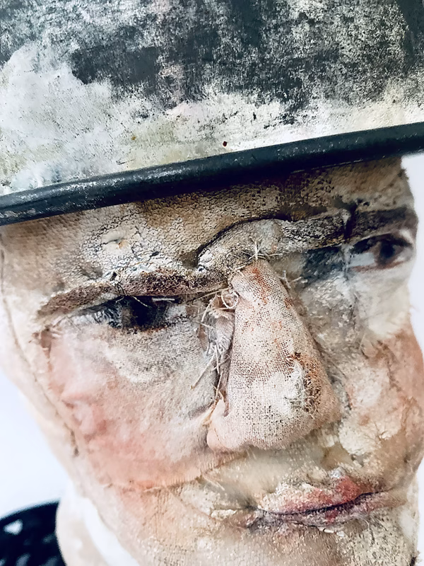 Close-up of head sculpture resting on a household iron trivet with a head piece of three stacked laboratory sieves.