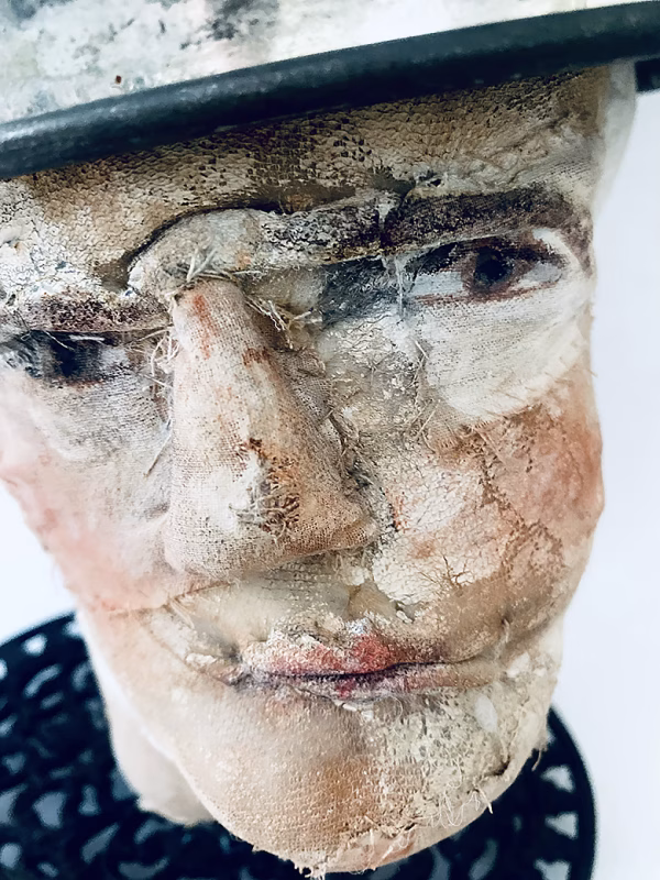 Close-up of head sculpture resting on a household iron trivet with a head piece of three stacked laboratory sieves.