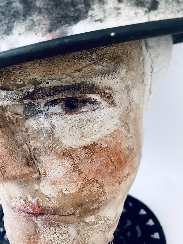 Close-up of head sculpture resting on a household iron trivet with a head piece of three stacked laboratory sieves.