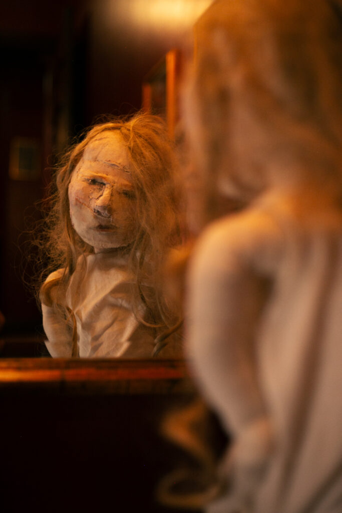 Puppet girl looking into a gold framed wall mirror in a theatre.